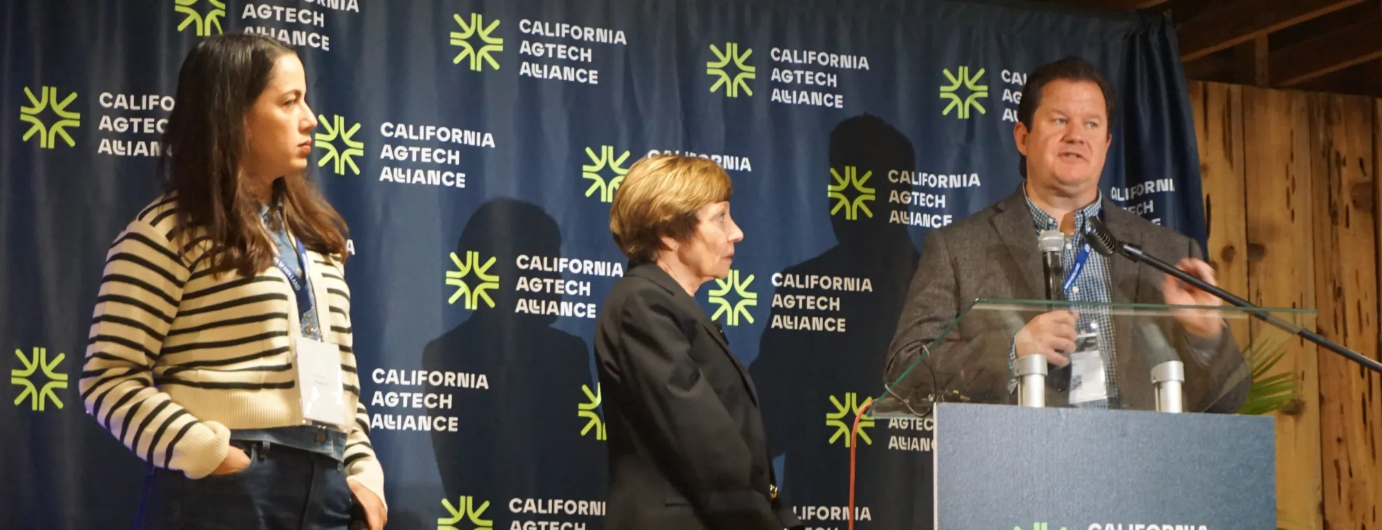 Gabe Youtsey speaks at a podium while Karen Aceves and Karen Ross look on at AgTech Alliance press conference