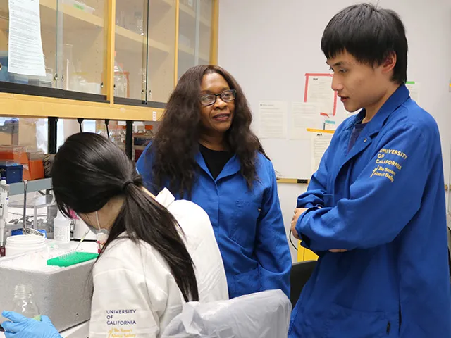 A woman sitting, a woman in lab coat speaks to a male student who is standing