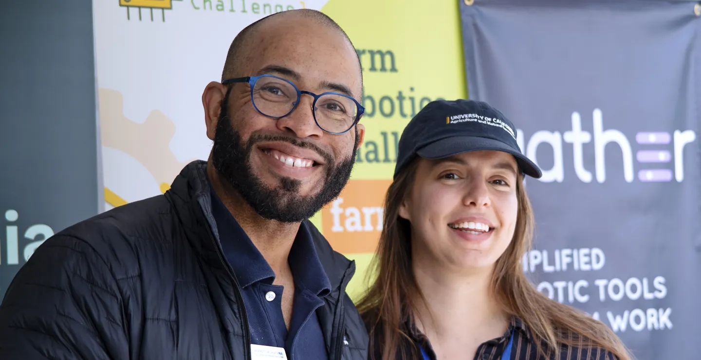 A man with glasses and woman in blue baseball cap smile in front of banners at Ag Day