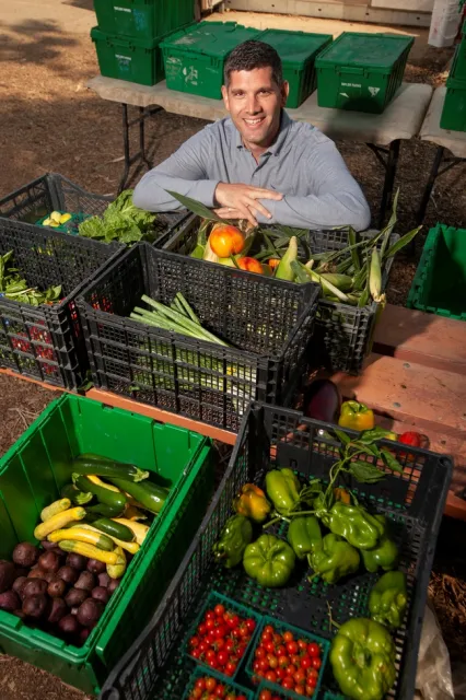 Ned Spang, surrounded by plastic bins of fresh produce, poses smiling with arms folded on edge of a bin containing apples and ears of corn.