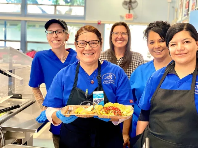 Vanessa Zajfen, wearing a hairnet and surrounded by four colleagues, holds a school lunch tray displaying a meal made with fresh produce.