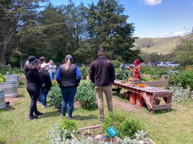 Henderson stands in a garden behind a table displaying flowers and plants as students listen