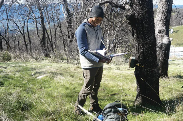 Surrounded by green grass, a man wearing knit cap and long-sleeved sweatshirt pages through a binder