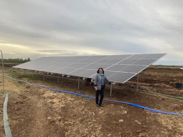 A woman stands next to an array of solar panels beside a fam field.