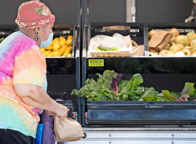 A woman looks at produce bins filled with leafy green lettuce, yellow squash and yellow onions.