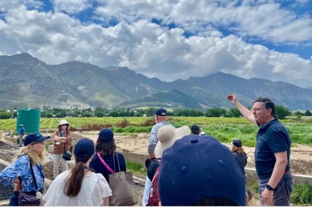 A man gestures toward a farm field as a group of people listen to him speak.