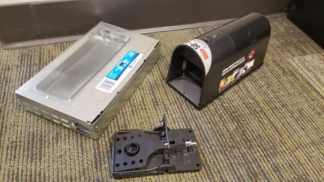 Three kinds of rodent traps displayed on a counter: one is a metal, rectangular box (an electrocution trap), one is a black box with a hinging door that is cylindrical on the top (a live-capture trap), and the third is a black plastic snapping trap with serrated teeth.