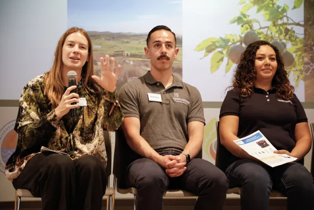 Three people sit on stage in front of a screen, speaking to an audience