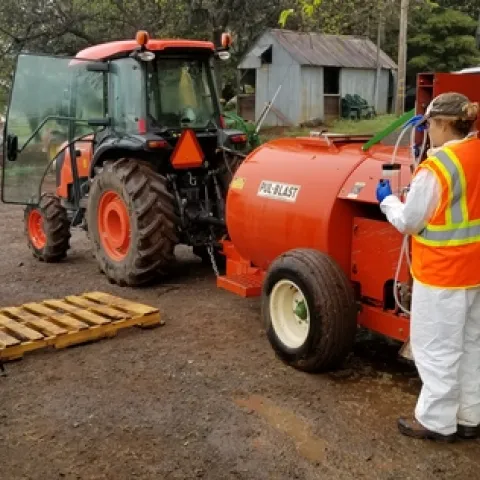 A photo showing a woman by an airblast sprayer being filmed while she measures flow rate from the nozzle.