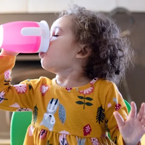 A toddler with curly brown hair drinks water from a pink sippy cup.