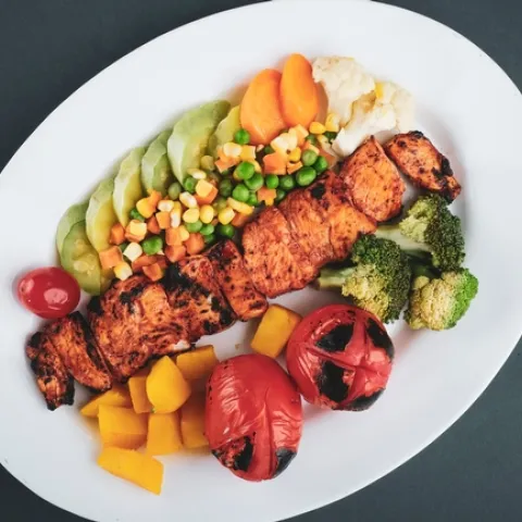 Grilled boneless chicken surrounded by broccoli, squash, corn, peas, carrots cauliflower and tomatoes on a white plate.