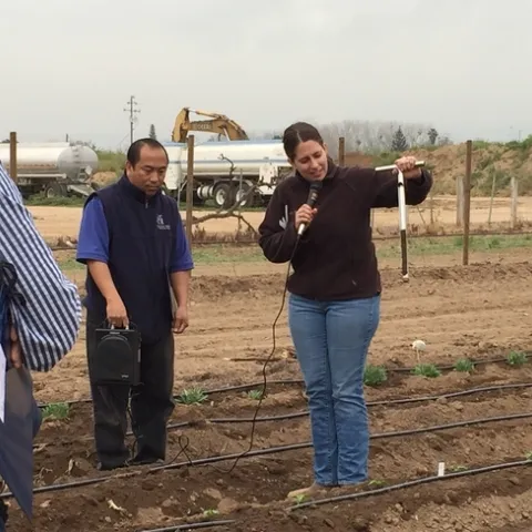 Dahlquist speaks into a mic while holding a soil sampler in a field lined with drip irrigation.