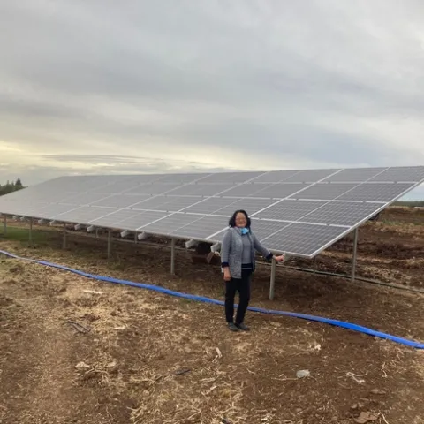 A woman stands next to an array of solar panels beside a fam field.
