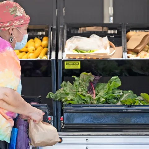 A woman looks at produce bins filled with leafy green lettuce, yellow squash and yellow onions.