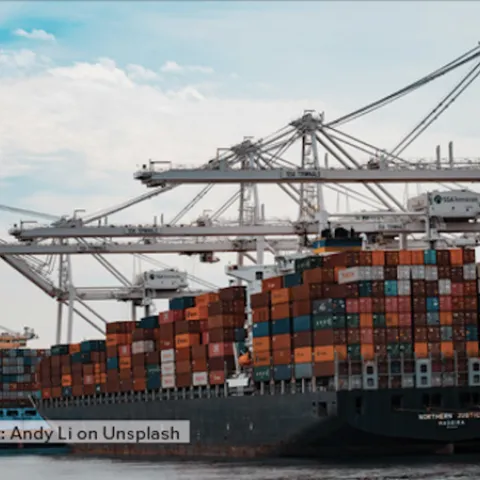 A ship loaded with shipping containers sits in port beneath cranes.