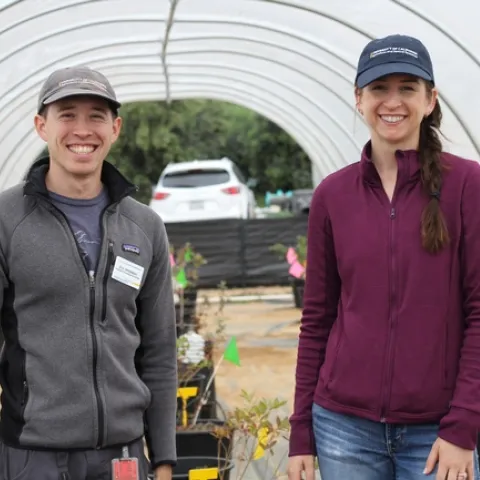 A young man and woman, both in their 30s, smile under a hoop house that is growing blueberries.