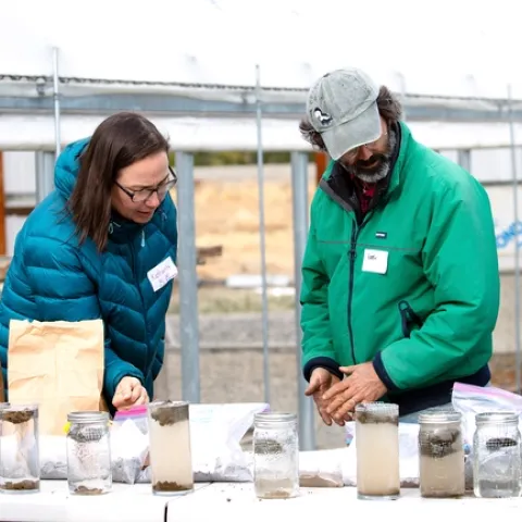 A woman in a puffy blue jacket and a bearded man in a green jacket look at slake test results in glass jars.