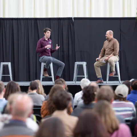 Swain sits on a stool on stage speaking to another man on stage