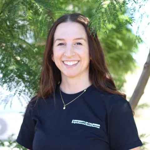 a photo of a woman with long brown hair wearing a black UC ANR branded t-shirt, standing in front of a tree