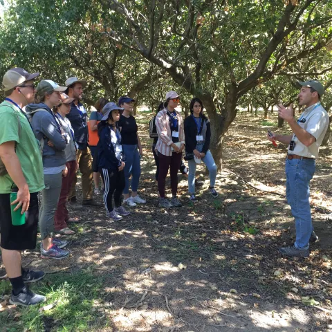 Houston Wilson, in baseball cap, gestures while doing a talk in the shade of an orchard