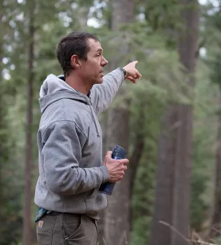 Rob York, holding a water bottle in his right hand, points into the forest with his left hand.