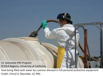 A person filling a tank with water while wearing chemical-resistant gloves, coveralls, chemical-resistant apron, and a face chield over safety glasses.