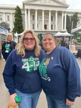 The image shows two women posing together, smiling. Both are wearing Sheldon 4-H tops and jeans.