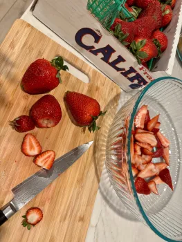 Box full of whole strawberries, a cutting board with partially-chopped strawberries, and a glass bowl full of chopped strawberries.