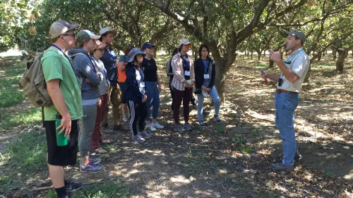 Houston Wilson, in baseball cap, gestures while doing a talk in the shade of an orchard