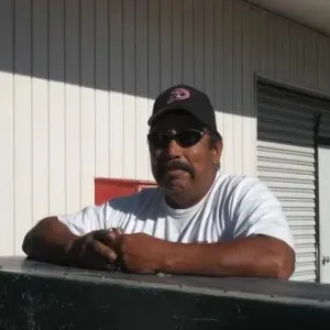 a man wearing a hat and sunglasses with his arms folded and torso leaning against the bed of a truck