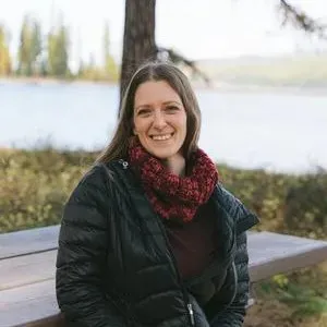 a woman with brown hair wearing a black puffer jacket and red scarf sitting at a picnic table near a body of water
