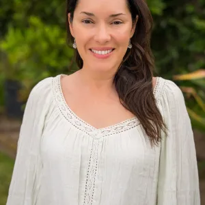 a woman with medium length brown hair wearing a white blouse standing in front of a beautiful green bush